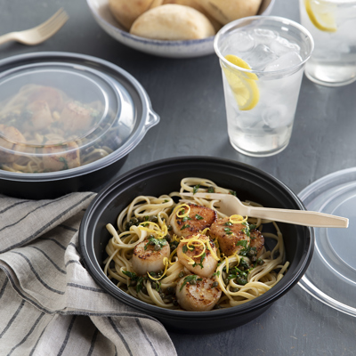 Black round take out container with pasta and grilled scallops inside