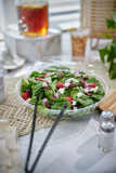 large salad in a clear catering container with black serving tongs	