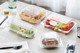 a sandwich, a wrap, sliced bell peppers, and cookies, all in different clear hinge lid takeout containers on a dining table