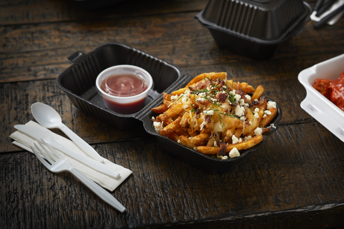 takeout container loaded fries with cheese, bacon, and chives with a red dipping sauce in a portion cup