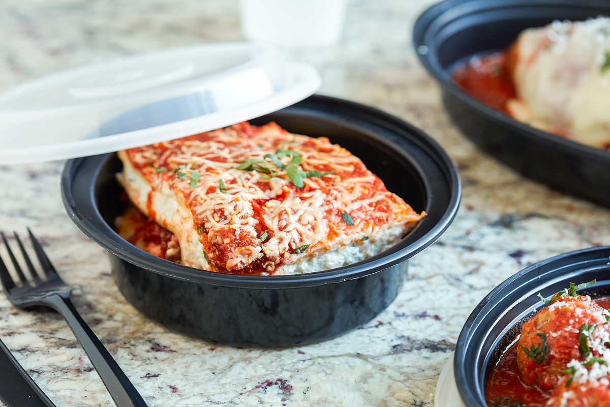 lasagna in a round black takeout container with clear lid	