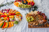 three different size clear catering platters one with fresh sliced veggies, one with fresh fruits, and one with nuts and crackers	