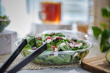 large salad in a clear catering container with black serving tongs