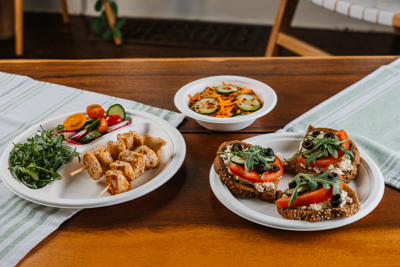 chicken kebabs, sliced veggies, cucumber and carrot salad, and vegetarian toast on different kinds of compostable plates	